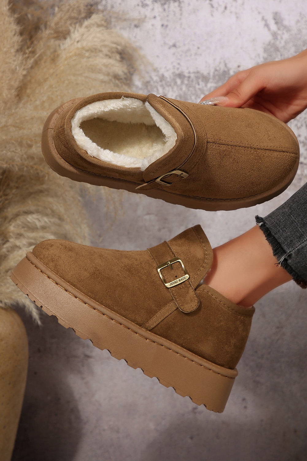 Brown suede shoes with buckles held by a hand against a textured background