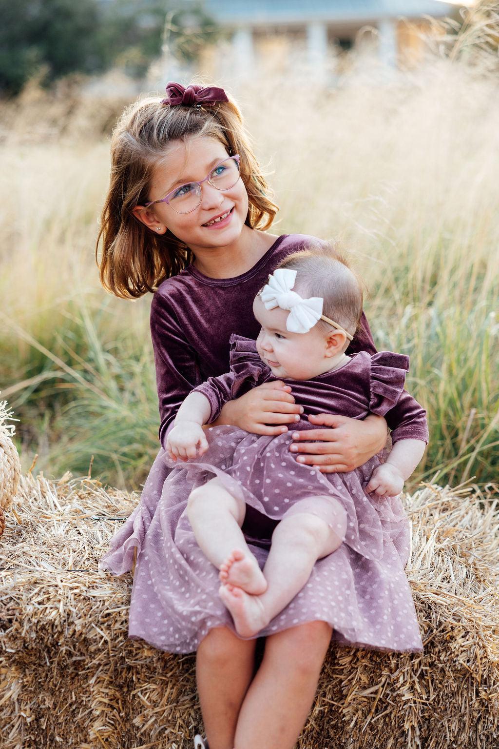 Two young girls in matching dresses sitting on a hay bale in a field.