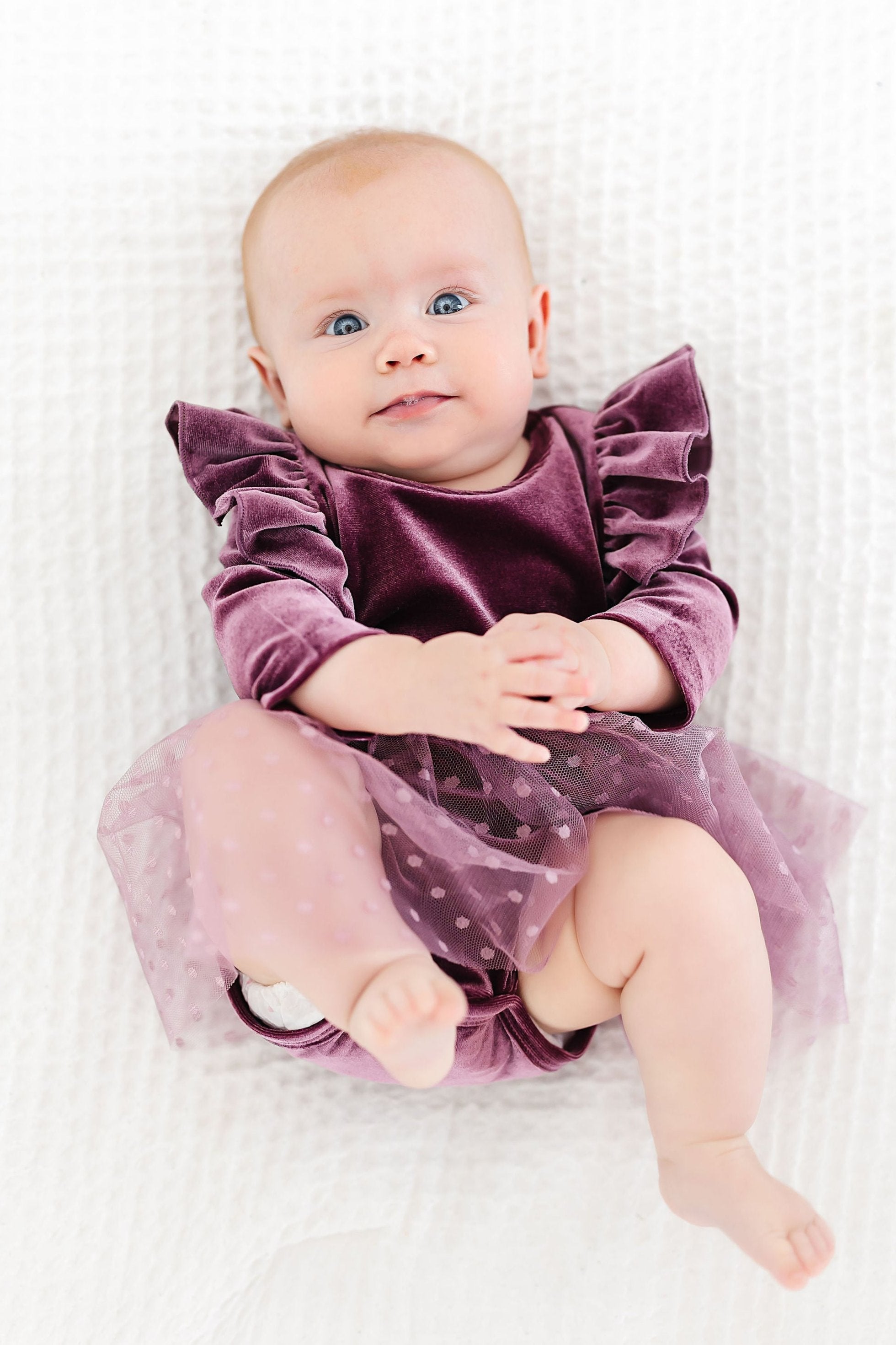Baby wearing a purple velvet dress with ruffles on a white blanket