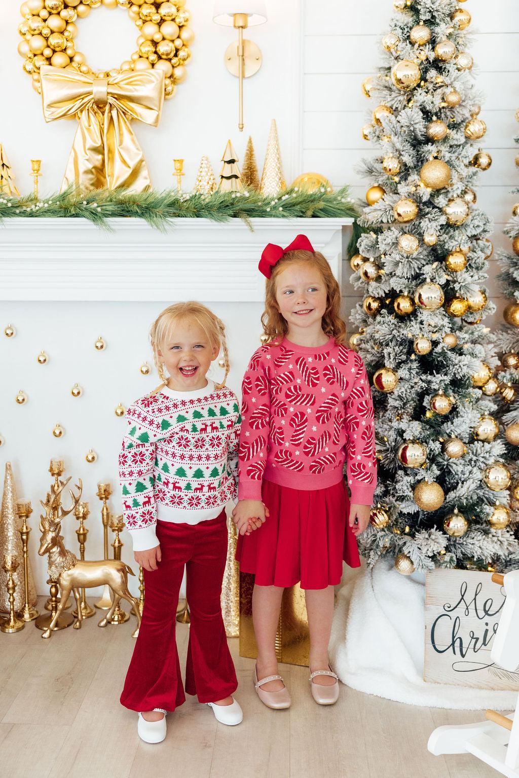 Two children in festive outfits standing in front of a decorated Christmas tree and fireplace.