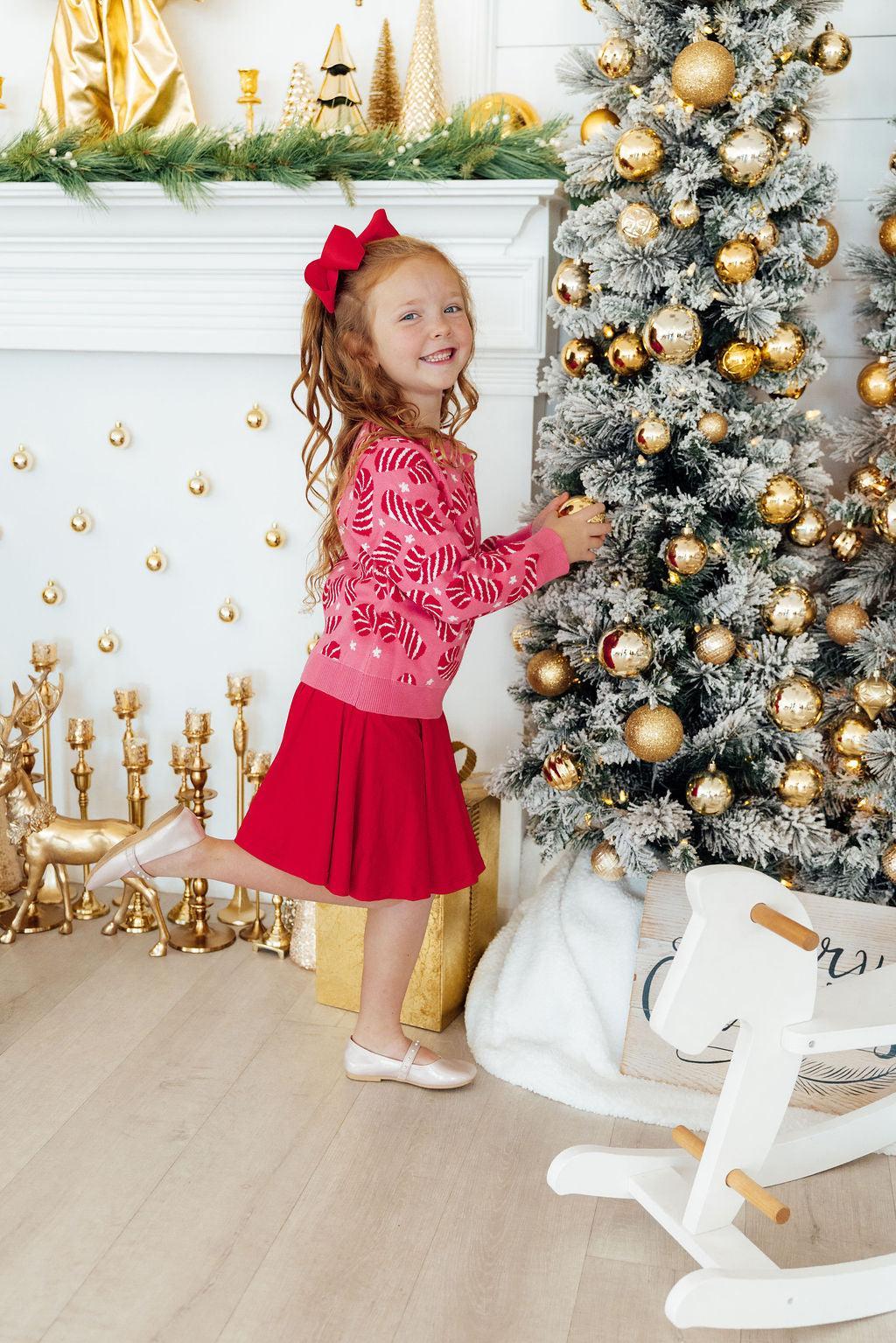 Child in a red dress standing next to a decorated Christmas tree in a festive room.