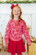 Young girl wearing a pink sweater with red patterns and a red skirt, standing in front of a decorated fireplace.