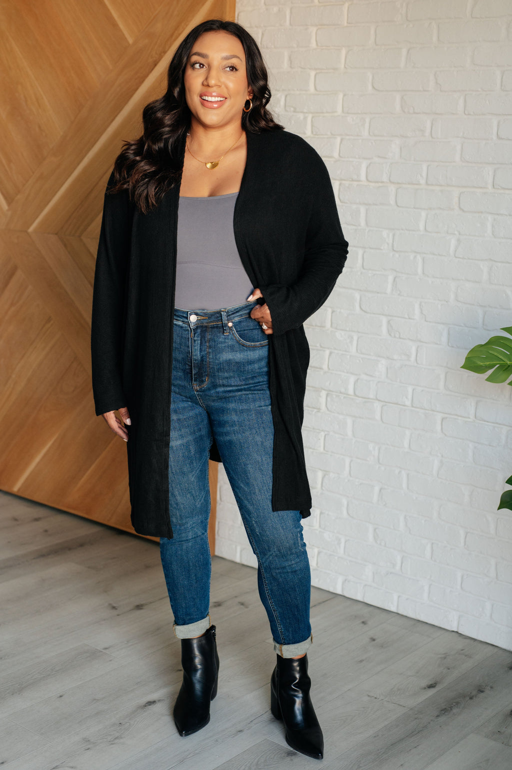 A woman modeling a long-line black cardigan over a gray ribbed top, paired with blue skinny jeans, rolled cuffs, and black ankle boots, standing in a brightly lit room with a wood-paneled wall and white wall.