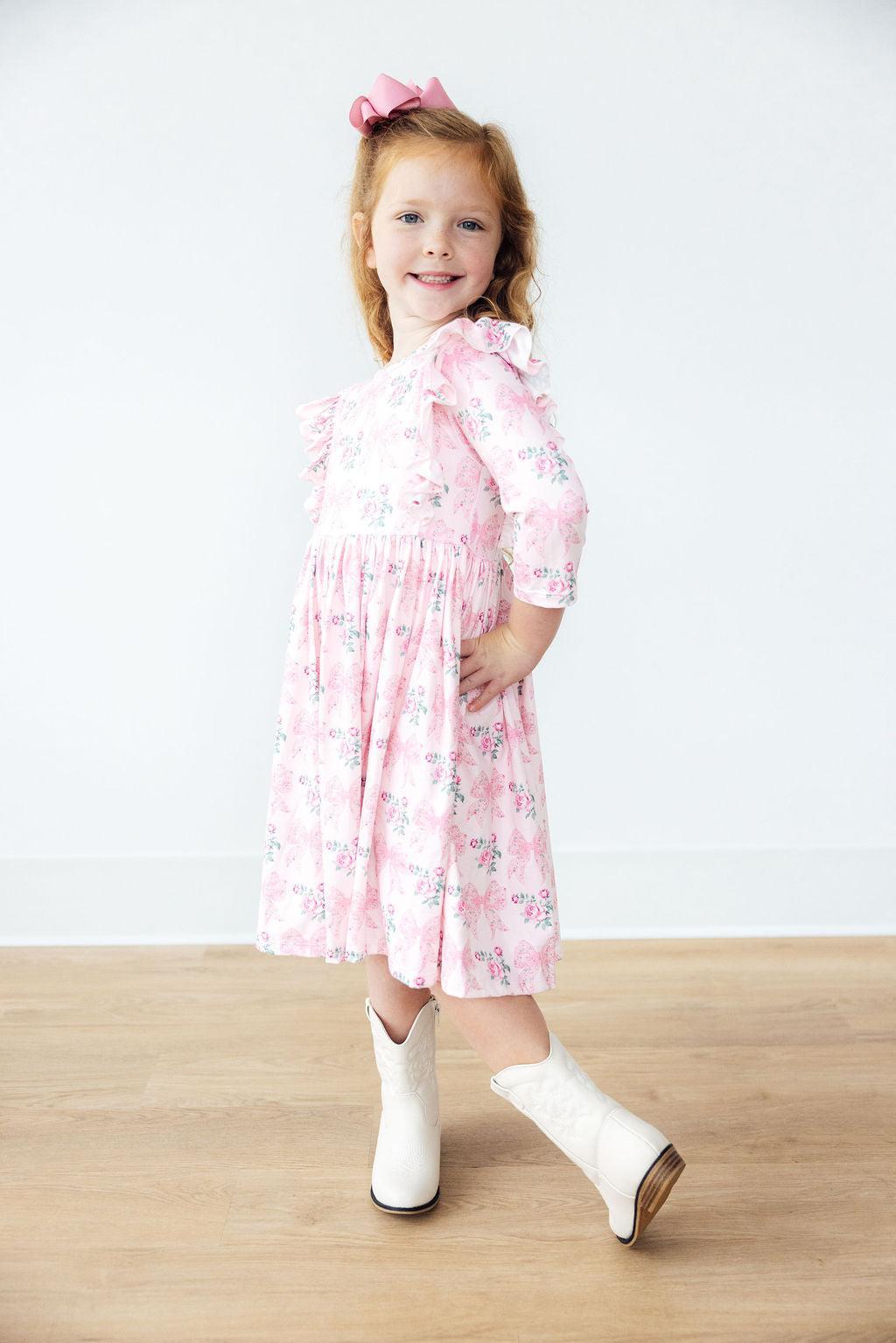 Young girl in a pink floral dress and white boots standing on a wooden floor with a white background