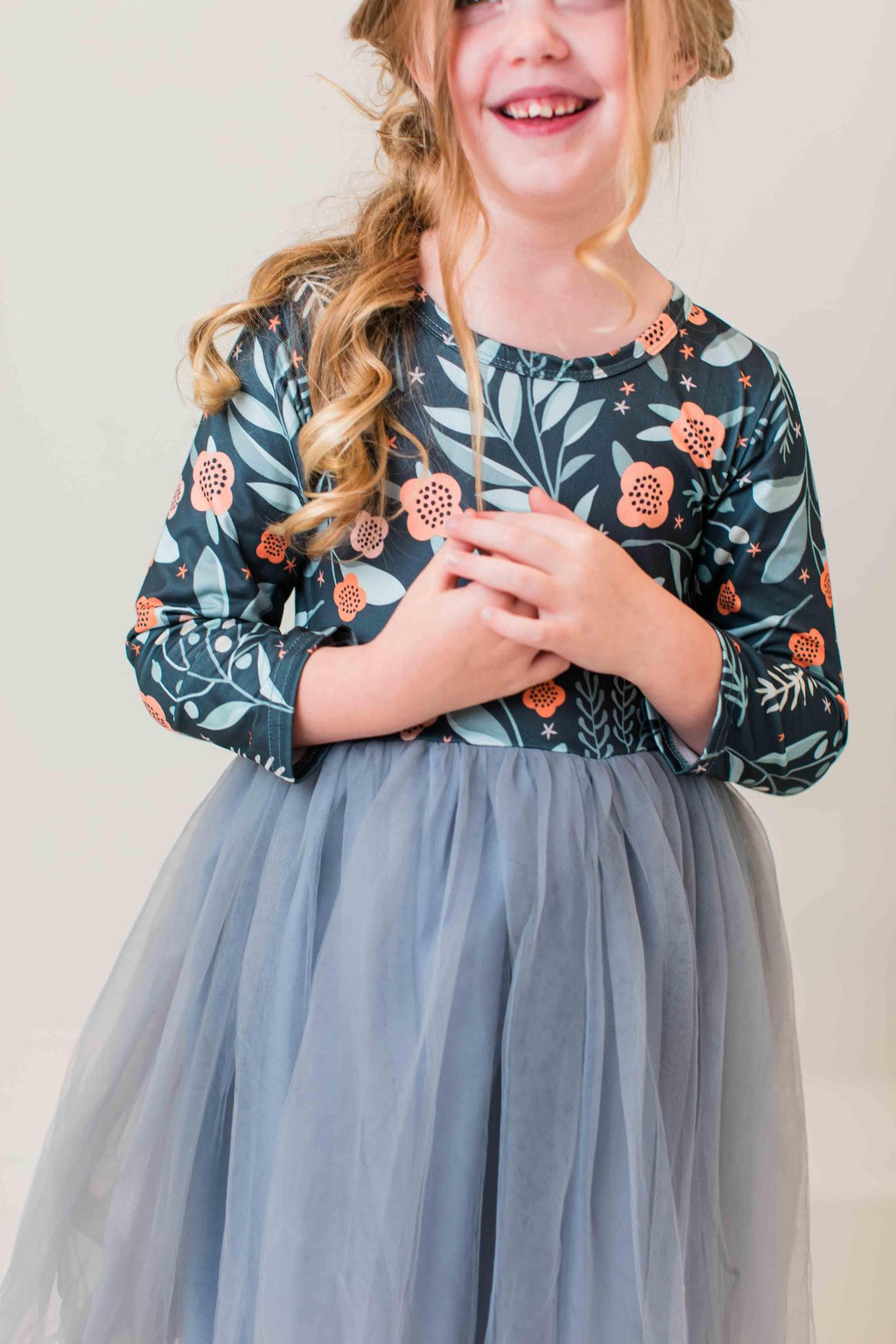 A young girl wearing a long-sleeve floral print top with a dusty blue tulle skirt, smiling against a plain light background.