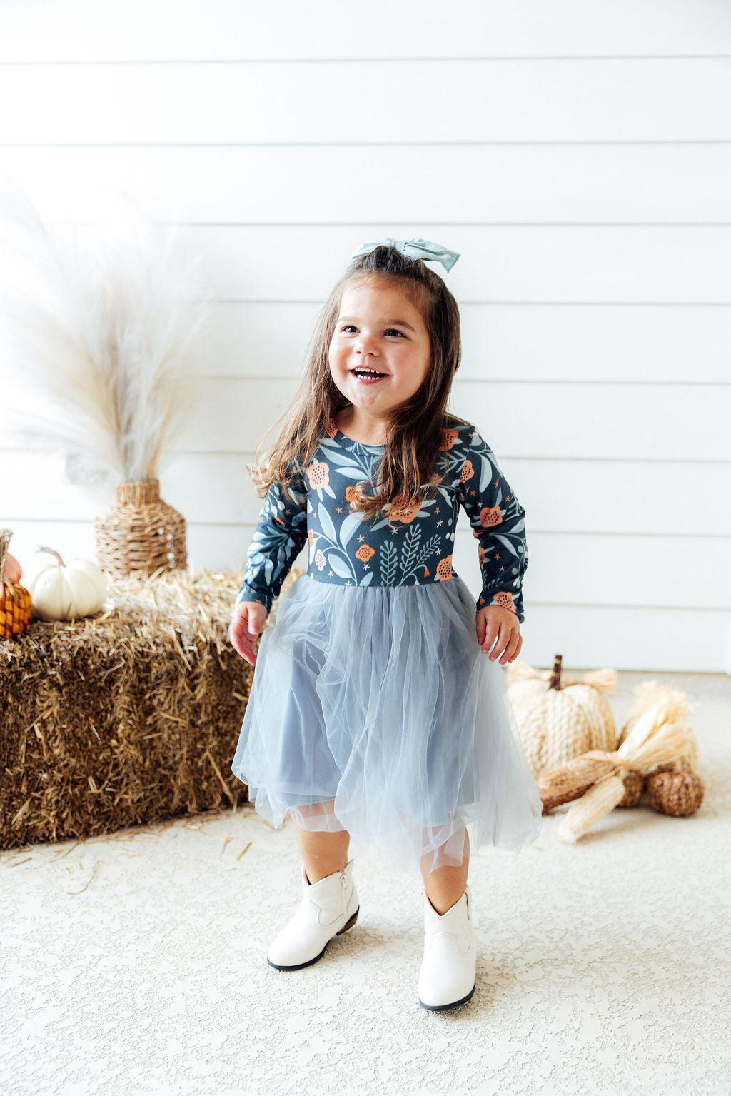 A young girl smiling and wearing a long-sleeve navy blue floral top with a light blue tulle skirt and white cowboy boots, standing in a styled room with hay bales and pumpkins.