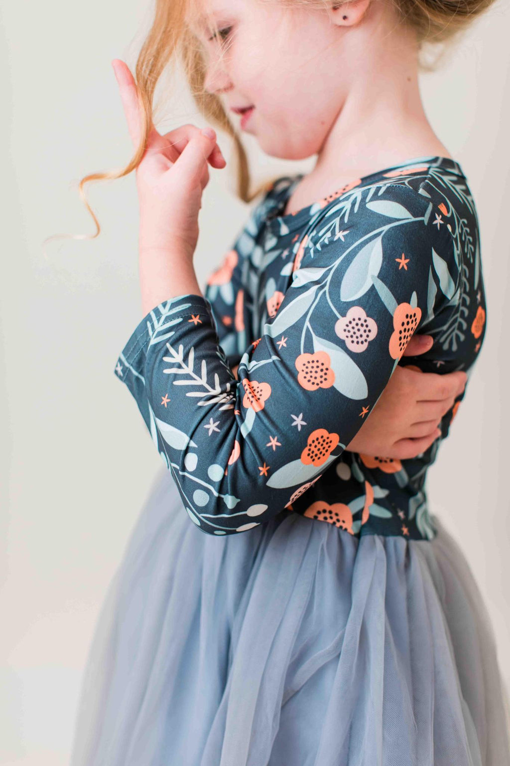 A young girl wearing a long-sleeve tulle dress with a navy blue floral bodice and a light blue-gray tulle skirt, shown from the side against a plain light background.
