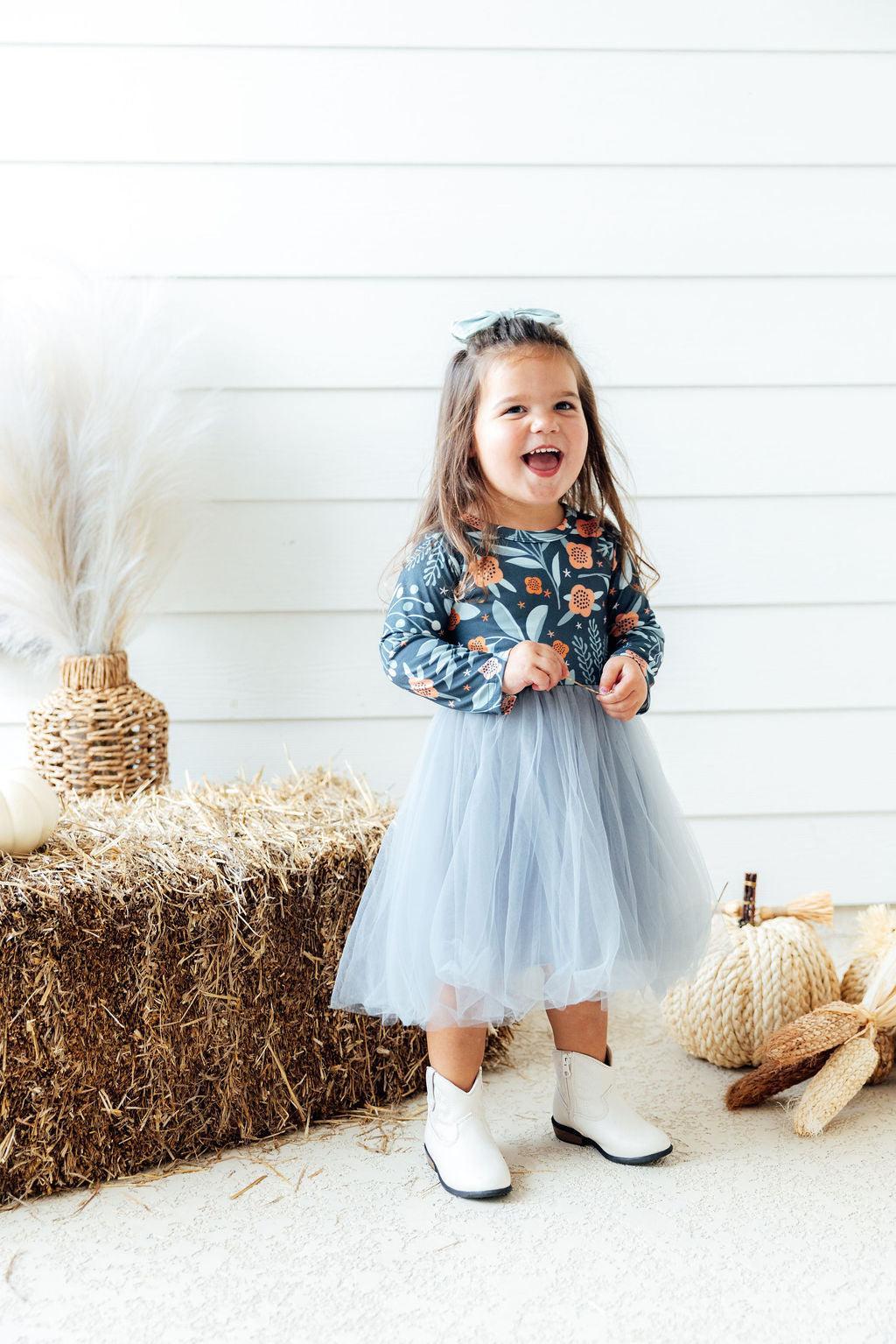 A young girl stands in a styled indoor autumn setting wearing a long-sleeve floral print top with a light blue tulle tutu skirt and white cowboy boots. Hay bale, pampas grass, and woven pumpkins are visible in the background.