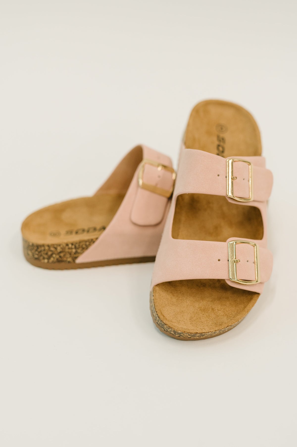 A pair of pink double-strap slide sandals with gold-tone buckles and cork soles, photographed on a clean white background.