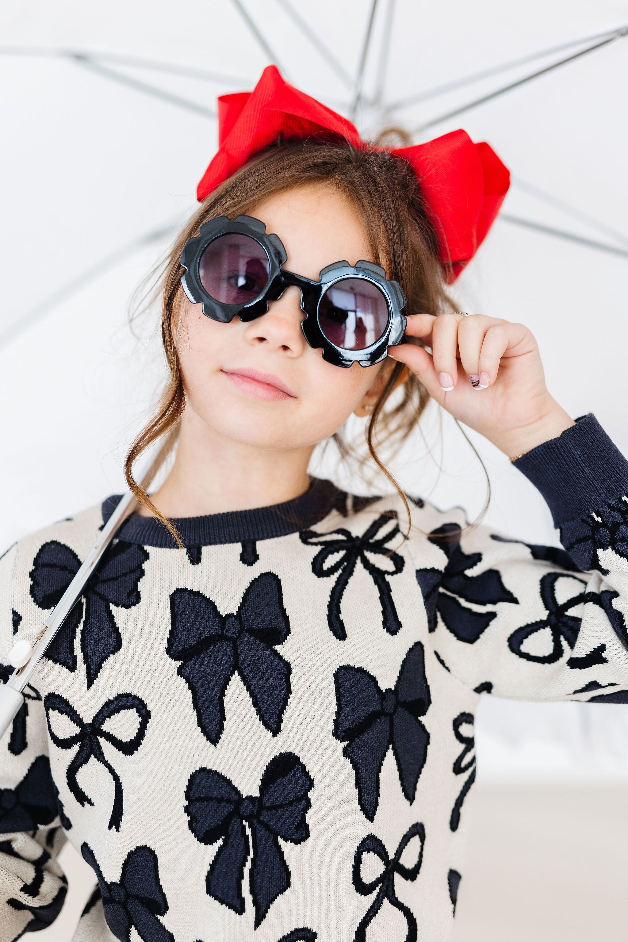 Child wearing a sweater with black bows, sunglasses, and a red bow headband.