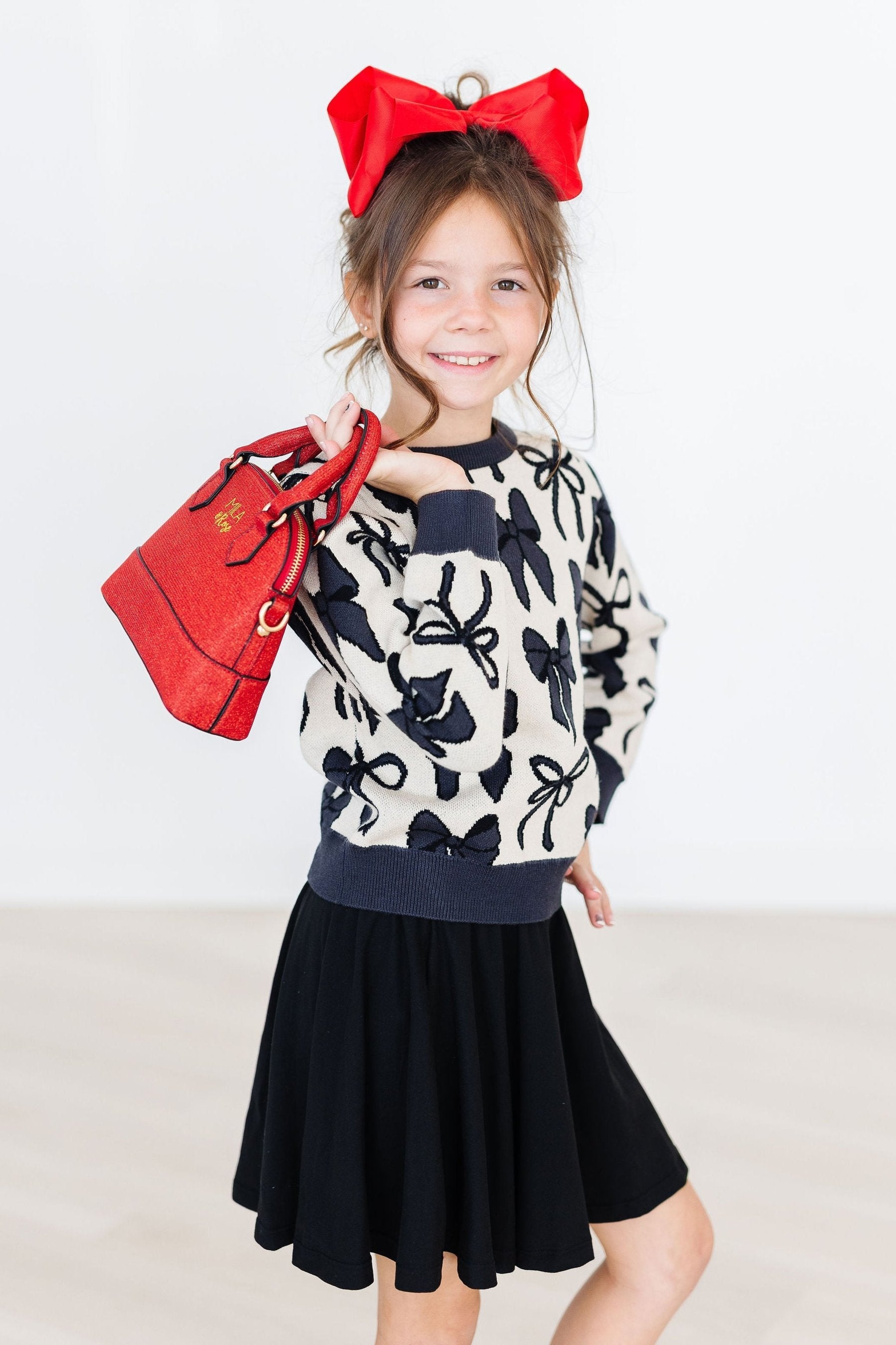 Young girl wearing a patterned sweater and black skirt with a red bow and bag.
