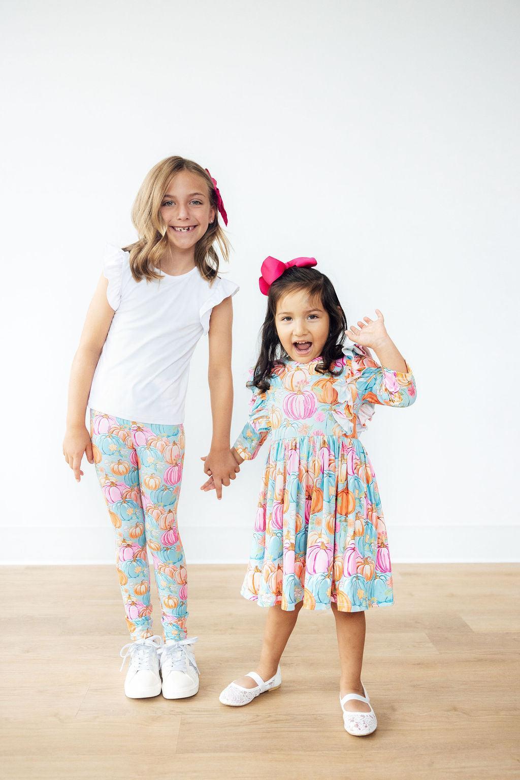 Two young girls holding hands, one in a white t-shirt and colorful pants, the other in a floral dress, on a wooden floor with a white background.