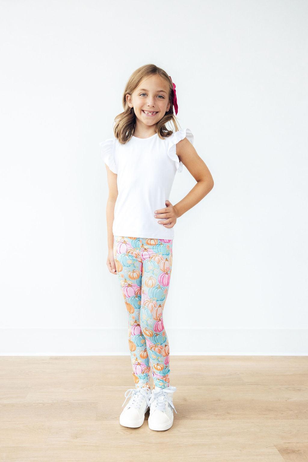 Young girl wearing a white t-shirt and patterned leggings standing on a wooden floor with a white background