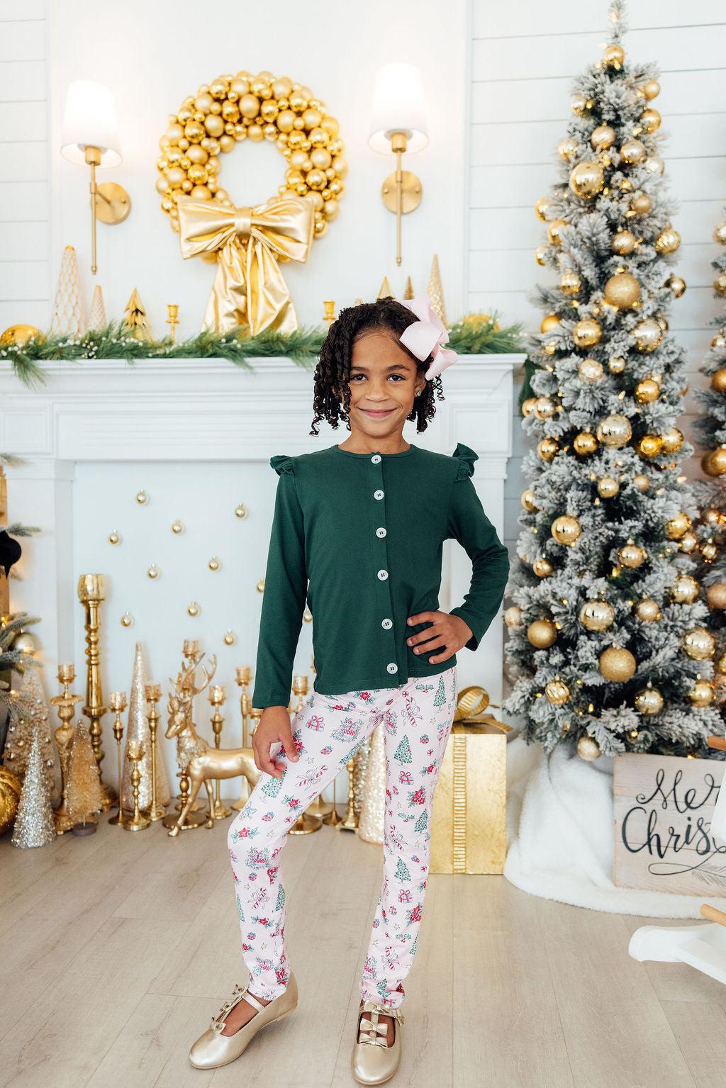 Young girl in festive outfit standing in front of a decorated Christmas tree and fireplace.