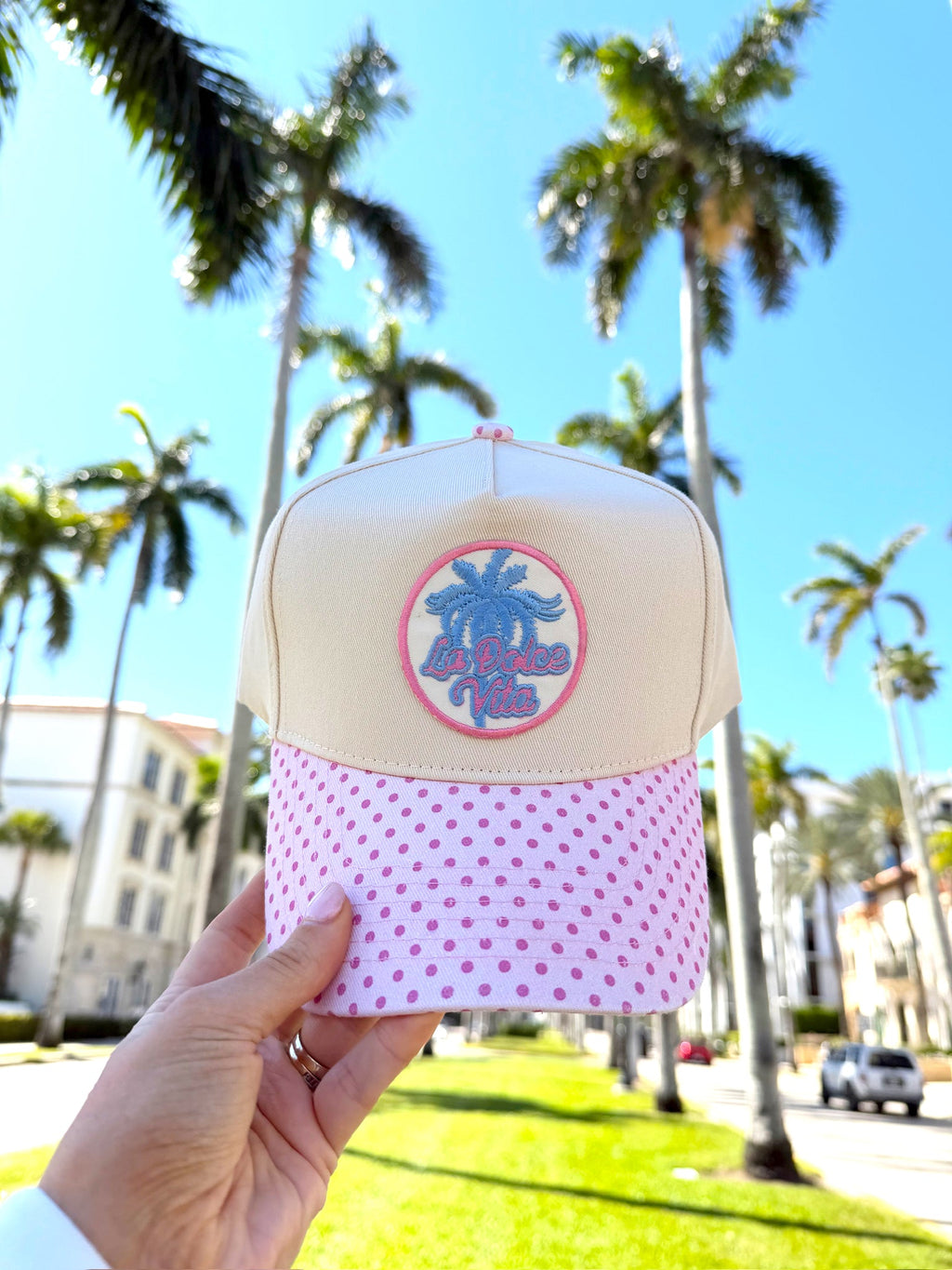 Beige and pink cap with palm tree design held in front of palm trees and buildings