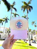 Beige and pink cap with palm tree design held in front of palm trees and buildings