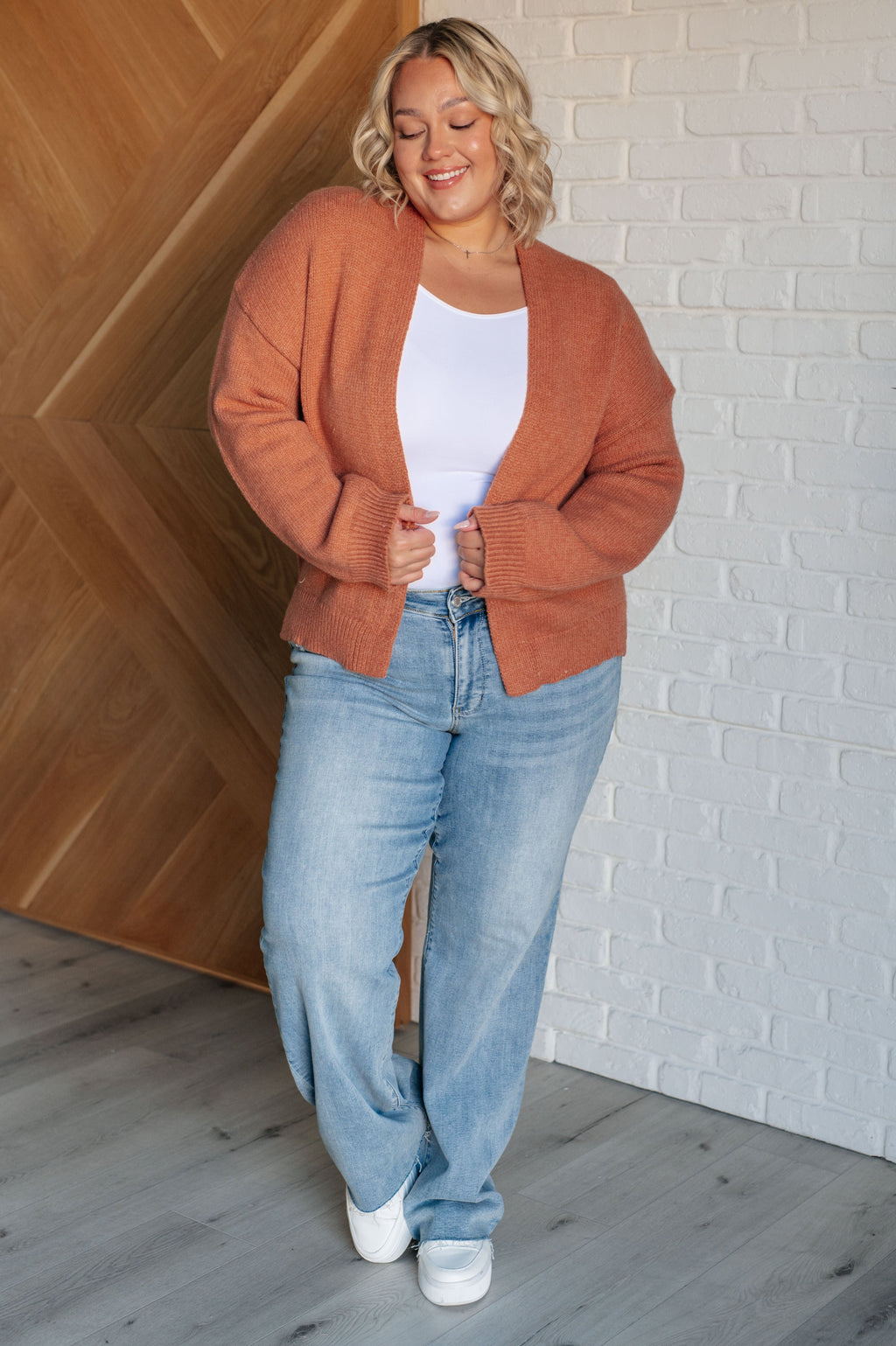 Woman wearing a rust-colored cardigan, white top, and blue jeans against a white brick wall.