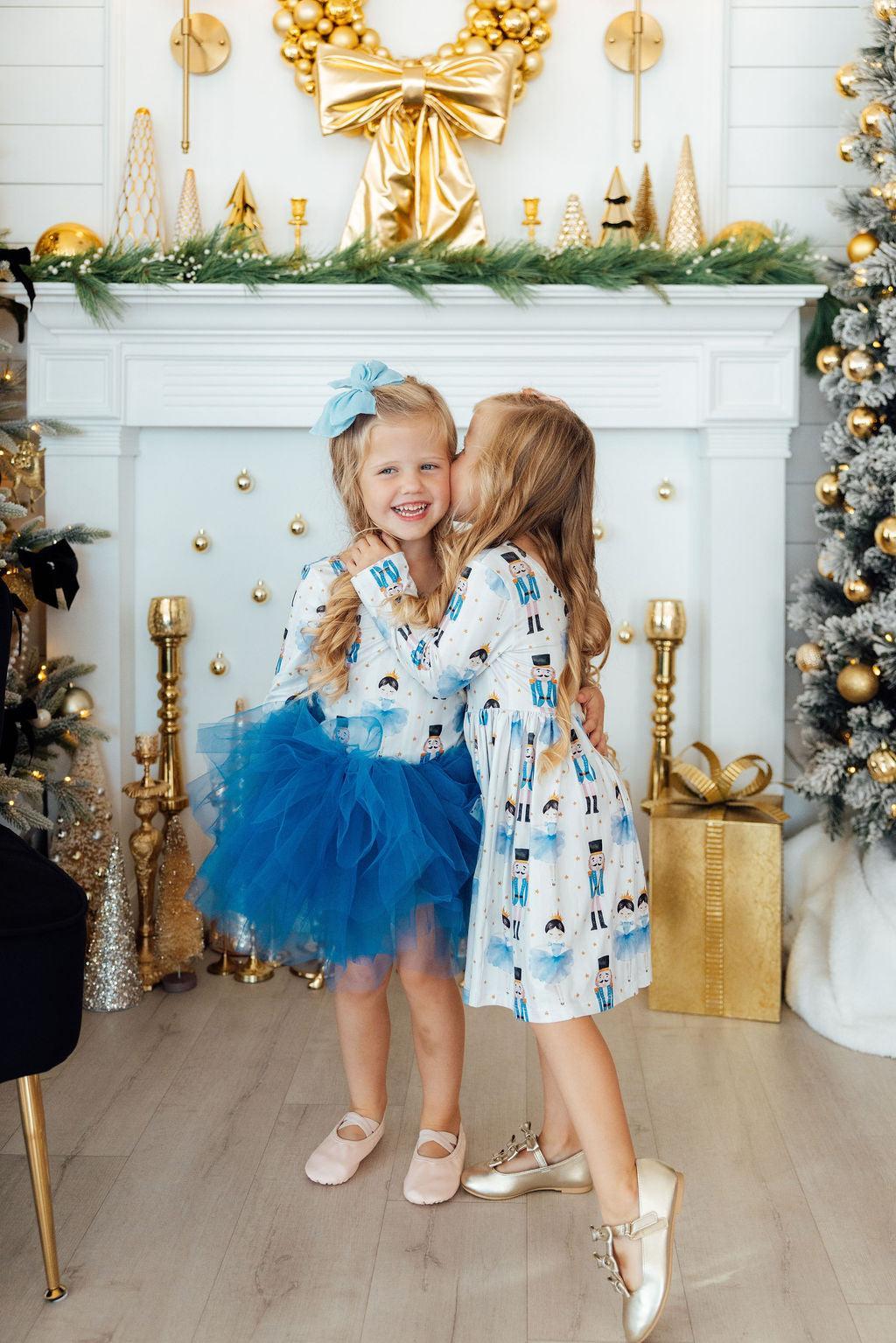 Two young girls in festive outfits standing in a decorated room with Christmas trees and a fireplace.