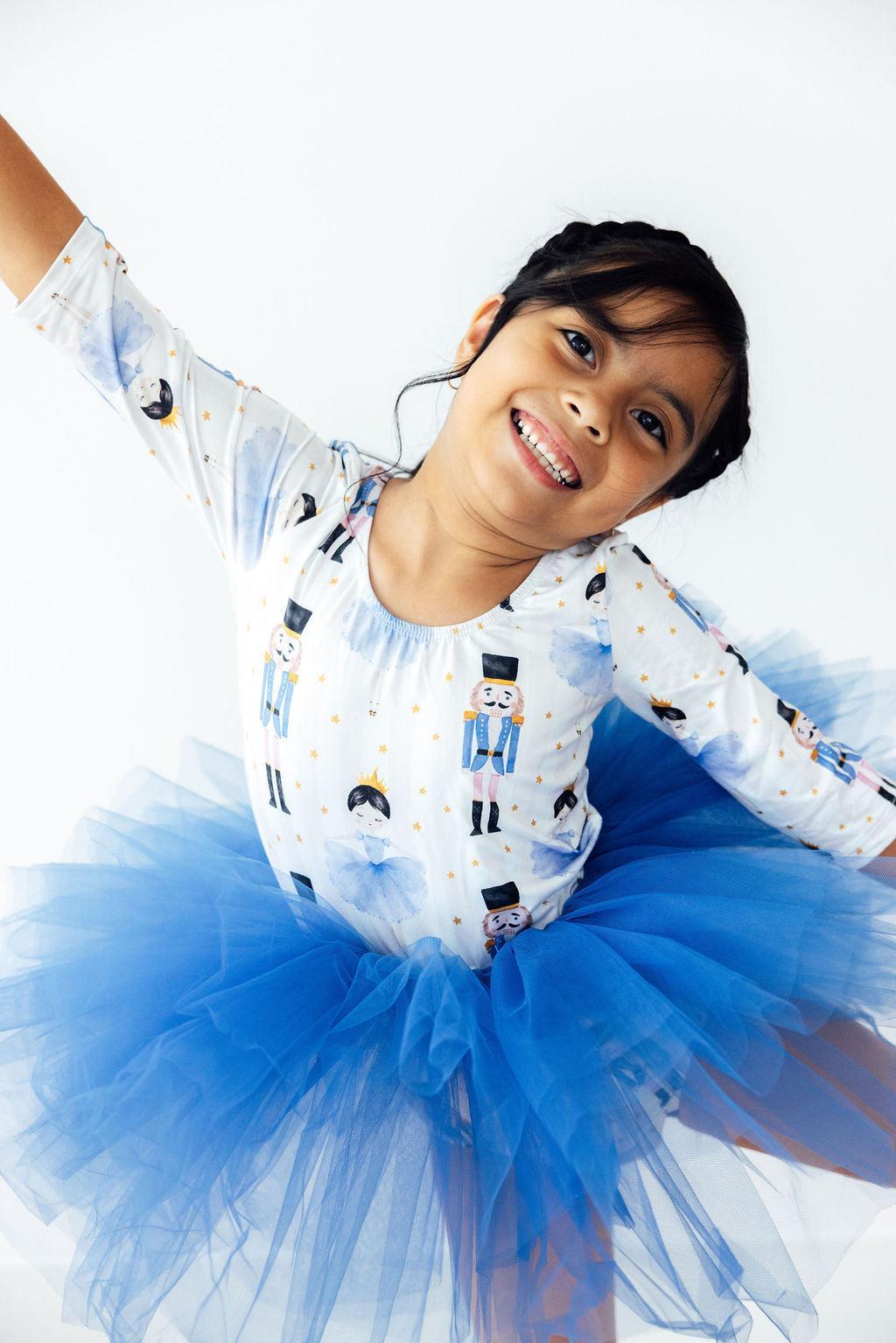 Child wearing a white long-sleeve shirt with black and blue designs and a blue tutu skirt on a white background