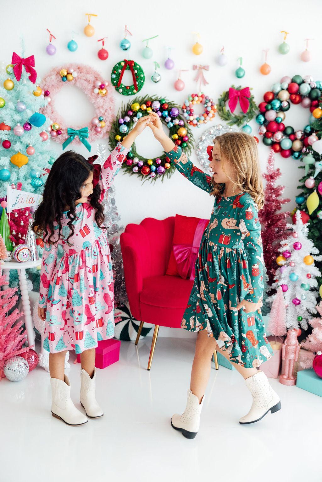 Two children in festive dresses standing in a decorated room with Christmas wreaths and ornaments.