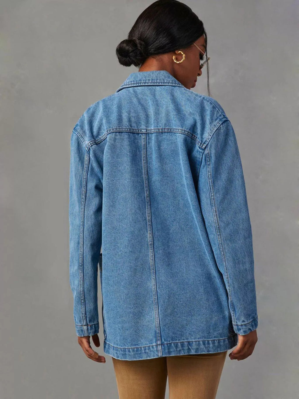 A woman shown from the back wearing an oversized light blue denim jacket, paired with tan leggings, against a textured gray studio background.