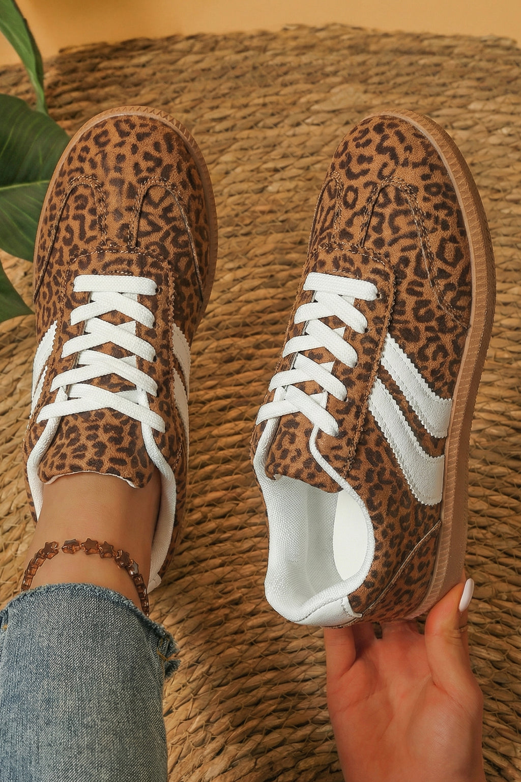 A pair of low-top sneakers with a brown and black leopard print upper, white laces, and white side stripes. One shoe is worn on a foot with blue jeans visible, and the other is held by a hand against a woven straw background.