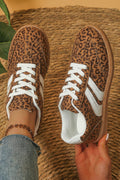 A pair of low-top sneakers with a brown and black leopard print upper, white laces, and white side stripes. One shoe is worn on a foot with blue jeans visible, and the other is held by a hand against a woven straw background.