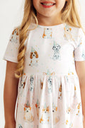 A close-up of a young girl wearing a short-sleeve white dress with a playful print of various dog breeds wearing party hats and small confetti dots.
