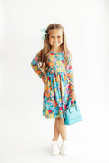 A young girl with long light brown hair wearing a colorful floral long-sleeve dress, a mint green hair bow, white sneakers, and holding a light blue handbag, standing against a plain white studio background.