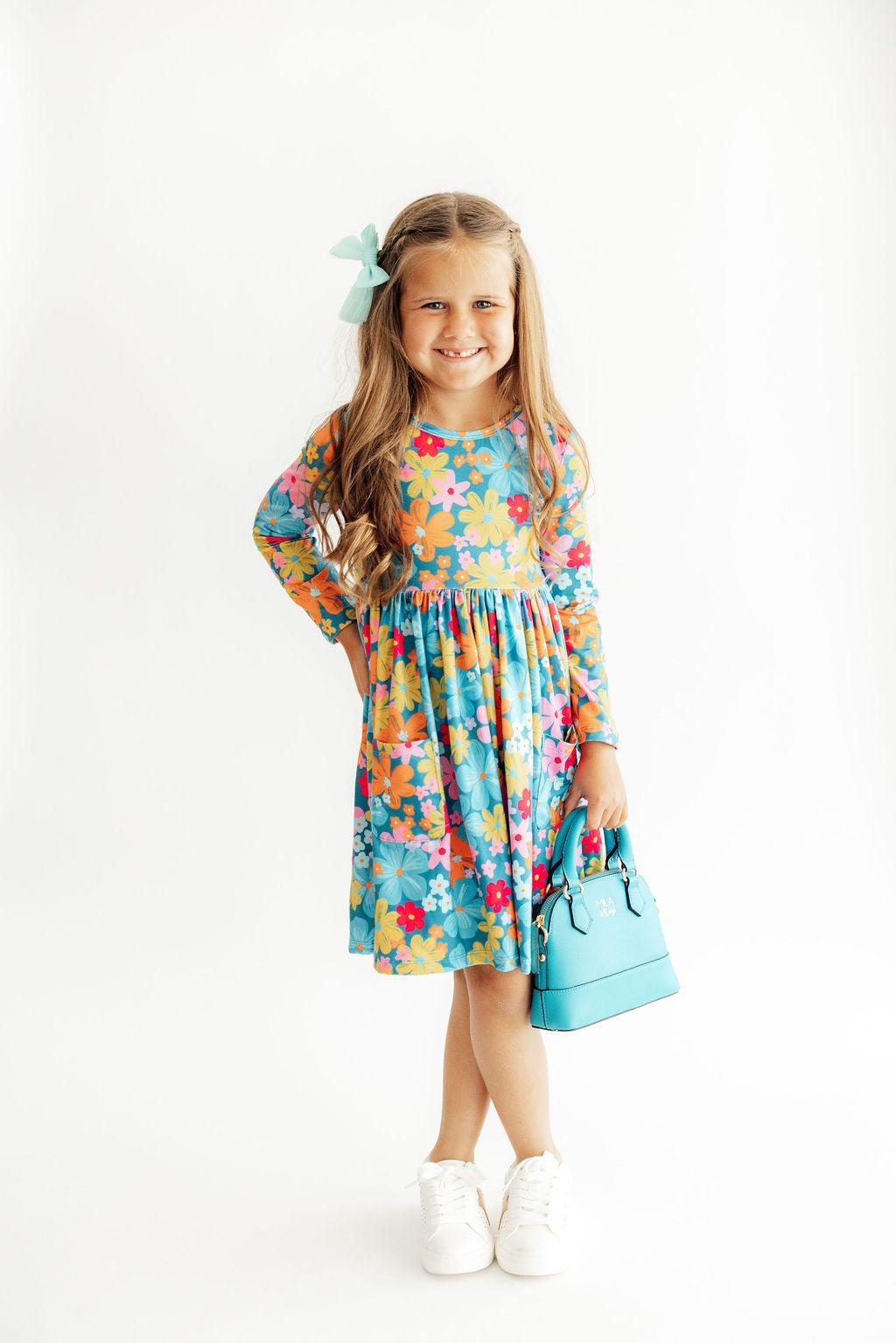 A young girl with long light brown hair wearing a colorful floral long-sleeve dress, a mint green hair bow, white sneakers, and holding a light blue handbag, standing against a plain white studio background.