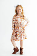 A young girl with red hair wearing a long-sleeved, knee-length dress with an autumn leaf pattern in orange, brown, and beige, paired with brown lace-up ankle boots and a small white bow headband, standing against a plain white background.