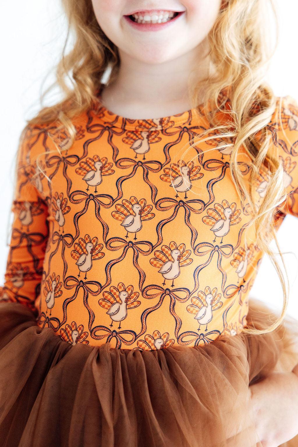 Child wearing an orange dress with a pattern of turkeys and bows, standing against a white background.